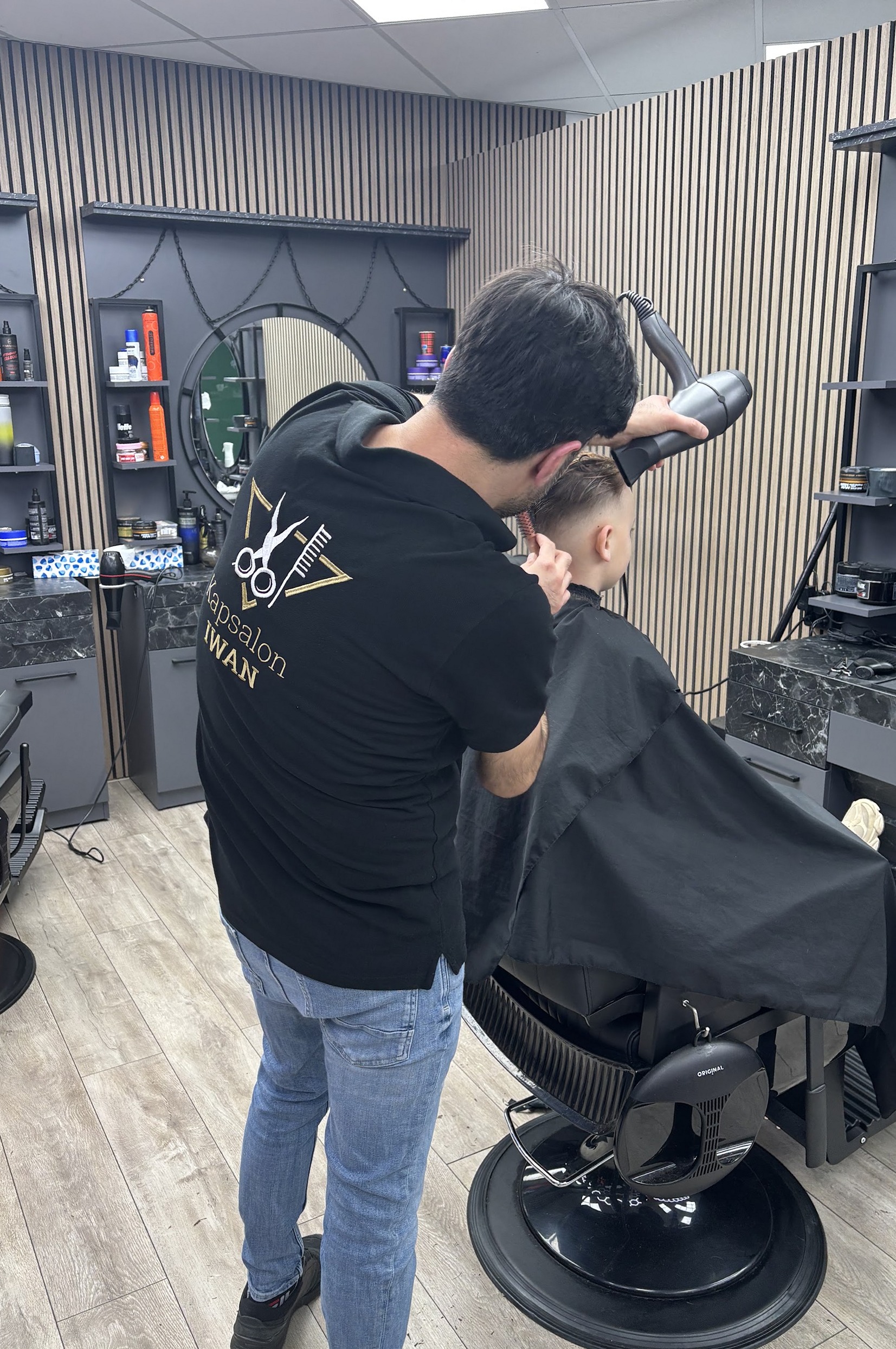 Een kapper in het zwarte Kapsalon IWAN-shirt föhnt de fade van een jongen in de kappersstoel