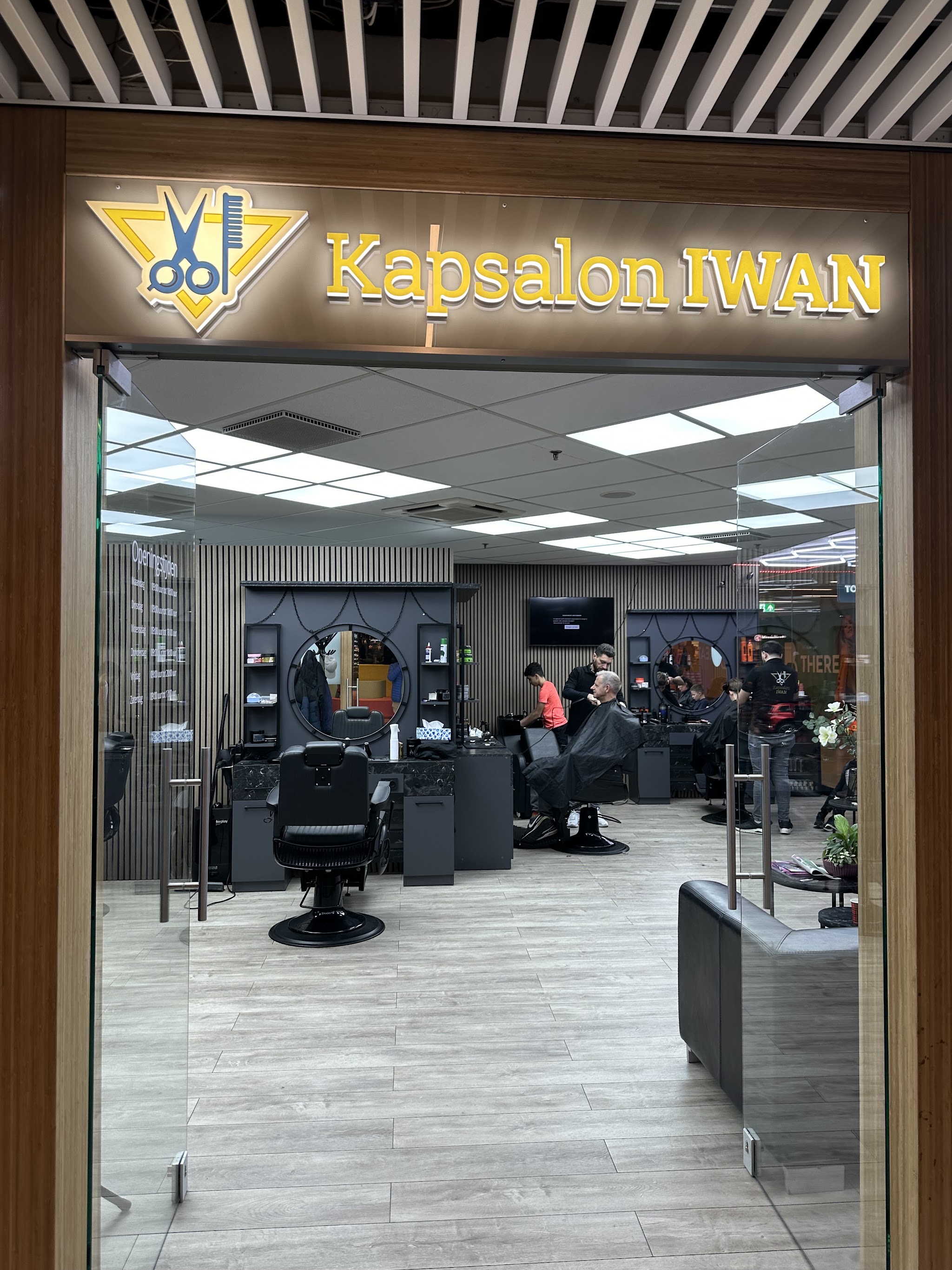 De ingang van Kapsalon Iwan in winkelcentrum Bisonspoor, met het gouden naambord boven de glazen deur en de kappersstoelen binnen